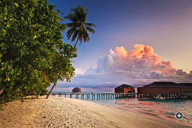 Dramatischer Sonnenaufgang am Strand mit Palmen und Wasserbungalows in Meerufenfushi, Malediven.