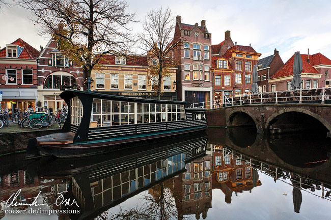 Ein Hausboot und mehrere Gebüude spiegeln sich in einem Kanal in Alkmaar, Niederlande.