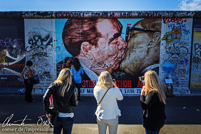 Drei Frauen betrachten auf der Berliner Mauer das Sozialistischer-Bruderkuss-Graffiti in Berlin, Deutschland.