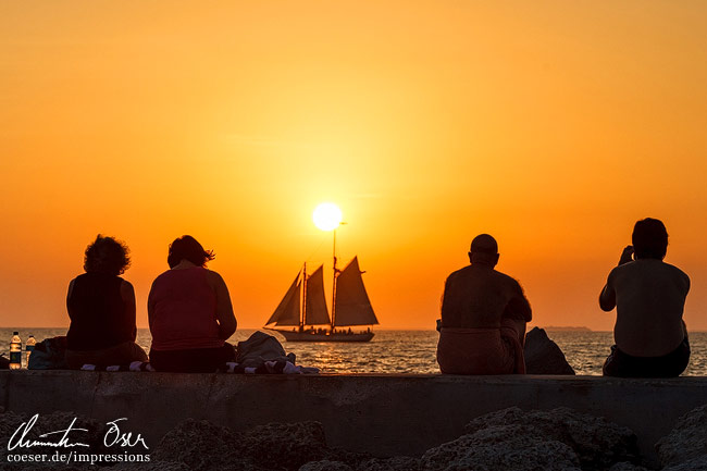 Menschen genießen den Sonnenuntergang und die Silhouette eines Segelschiffs am Zachary-Taylor-Strand in Key West, USA.