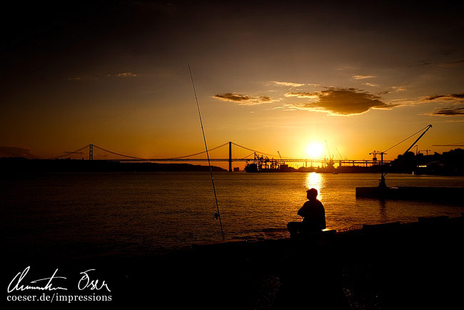 Die Silhouette eines Fischers im Sonnenuntergang vor der Ponte 25 de Abril Brücke in Lissabon, Portugal.