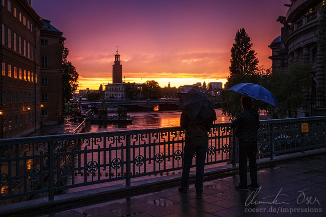 Zwei Personen blicken auf das Stadshus (Rathaus) und einen spektakulären Sonnenuntergang in Stockholm, Schweden.